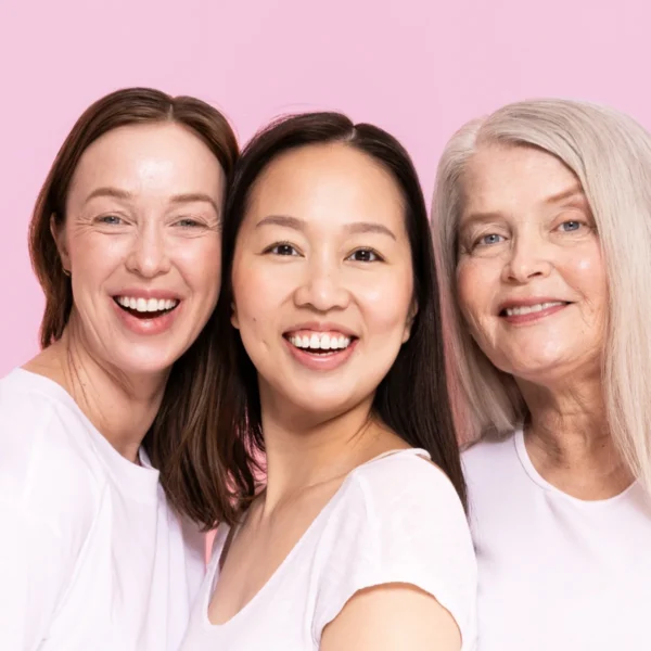 Three women of different ages smiling together against a pink background, conveying friendship and happiness.