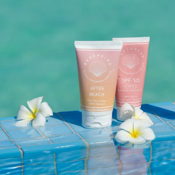 Two pink sunscreen tubes labeled SPF 50 and After Beach, resting on blue pool tiles with white plumeria flowers nearby.