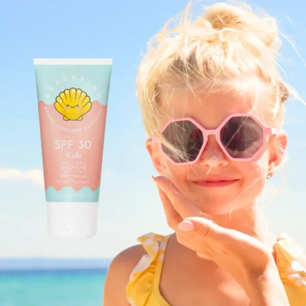Tube of SPF 30 Kids sunscreen with a yellow shell logo floats beside a smiling girl at the beach wearing pink heart-shaped sunglasses.