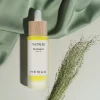 Bottle of Henua The Pause Bio-Retinol Serum with a wooden cap and yellow liquid, set against green fabric and dried grasses.