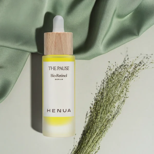 Bottle of Henua The Pause Bio-Retinol Serum with a wooden cap and yellow liquid, set against green fabric and dried grasses.