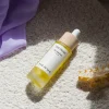 Bottle of Henua The Pause Bio-Retinol serum with a wooden dropper cap resting on a cream textured rug, near a purple fabric piece and a yellow hair clip nearby.