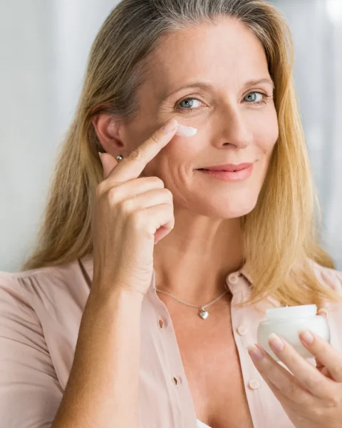 Woman applying moisturizer under her eye with her finger, holding a jar of cream.