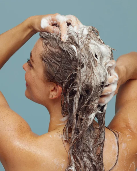 Person washing long hair with soap lather, seen from behind against a blue background, hands massaging the scalp.