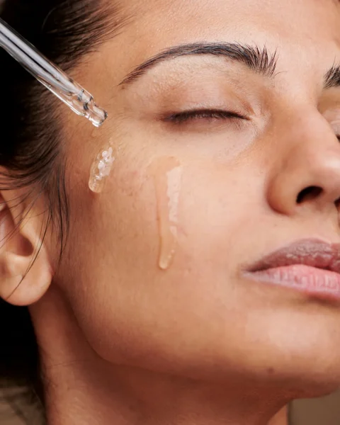 A close-up of a woman with closed eyes as a dropper releases serum onto her cheek for skincare.
