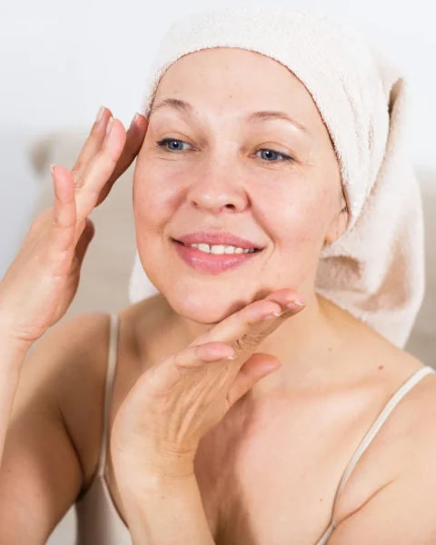 Woman with a towel wrapped around her head, gently touching her face and smiling in a skincare routine setting.