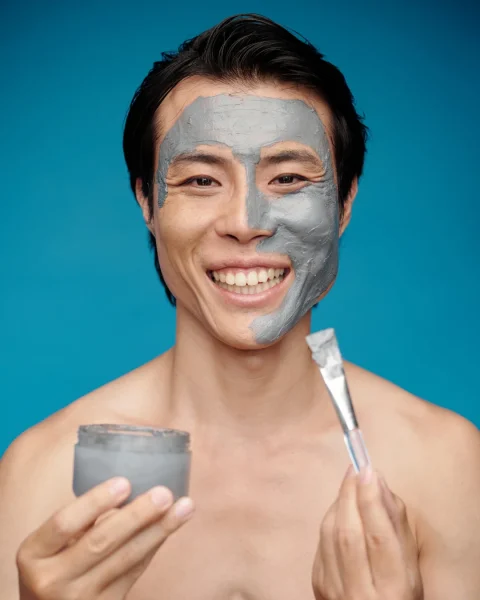Person smiling with a gray facial mask on their face, holding a jar of mask and a spatula against a blue background