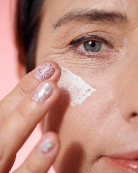 Close-up of a person removing under-eye makeup with a wipe, showing glittery nails and a pink backdrop.