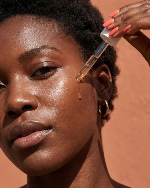 Close-up of a dark-skinned woman using a dropper to apply serum to her cheek; a few drops fall, with warm background.