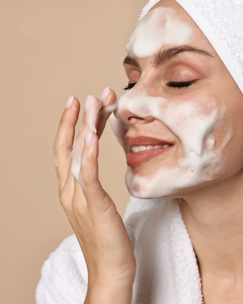 Woman with a foamy facial cleanser on her face, eyes closed and smiling, wrapped in a white towel.