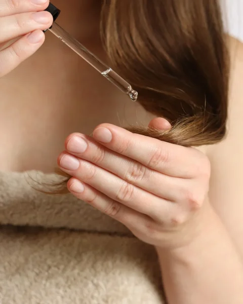 Close-up of a hand holding a dropper above a strand of brown hair, about to apply a drop of oil to the hair
