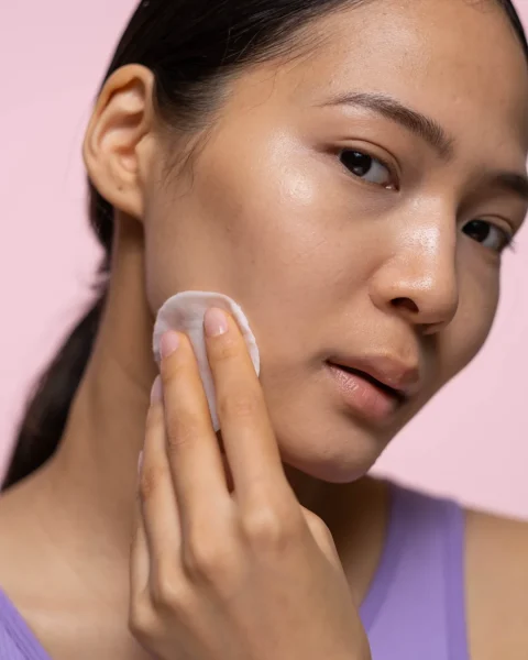 Close-up of a woman in a lavender top patting her cheek with a cotton pad against a pink background.
