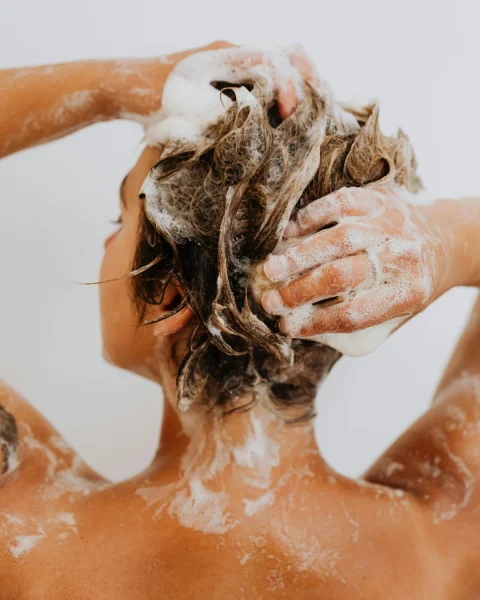 Back view of a person washing hair, hands massaging shampoo into wet brown hair with bubbles on shoulders and neck.